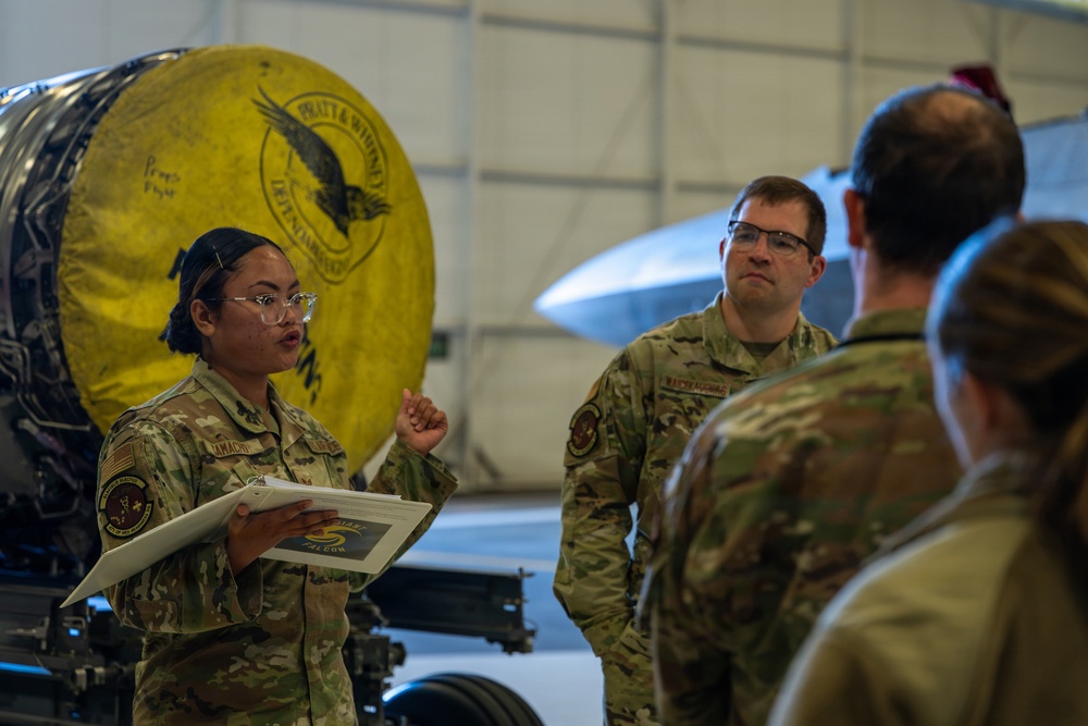Military personnel in camouflage uniforms are gathered inside an aircraft hangar for what appears to be a briefing or training session. A female service member wearing glasses and a name tape reading 'CAMACHO' is holding documents and speaking to the group. In the background, a large Pratt &amp; Whitney jet engine with a distinctive yellow cover is visible alongside what appears to be a military aircraft.