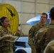 Military personnel in camouflage uniforms are gathered inside an aircraft hangar for what appears to be a briefing or training session. A female service member wearing glasses and a name tape reading 'CAMACHO' is holding documents and speaking to the group. In the background, a large Pratt &amp; Whitney jet engine with a distinctive yellow cover is visible alongside what appears to be a military aircraft.