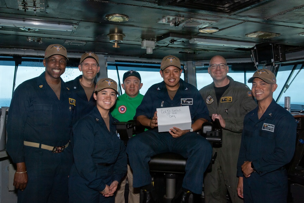 A group of eight individuals, mostly in Navy blue uniforms with tan caps, pose for a photo on what appears to be the bridge of a naval vessel. One person in the center is seated and holding a white sign that reads 'Gold Eagle of the Day.' One individual stands out wearing a green t-shirt with a Captain America shield logo, likely a civilian visitor or guest.