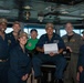 A group of eight individuals, mostly in Navy blue uniforms with tan caps, pose for a photo on what appears to be the bridge of a naval vessel. One person in the center is seated and holding a white sign that reads 'Gold Eagle of the Day.' One individual stands out wearing a green t-shirt with a Captain America shield logo, likely a civilian visitor or guest.