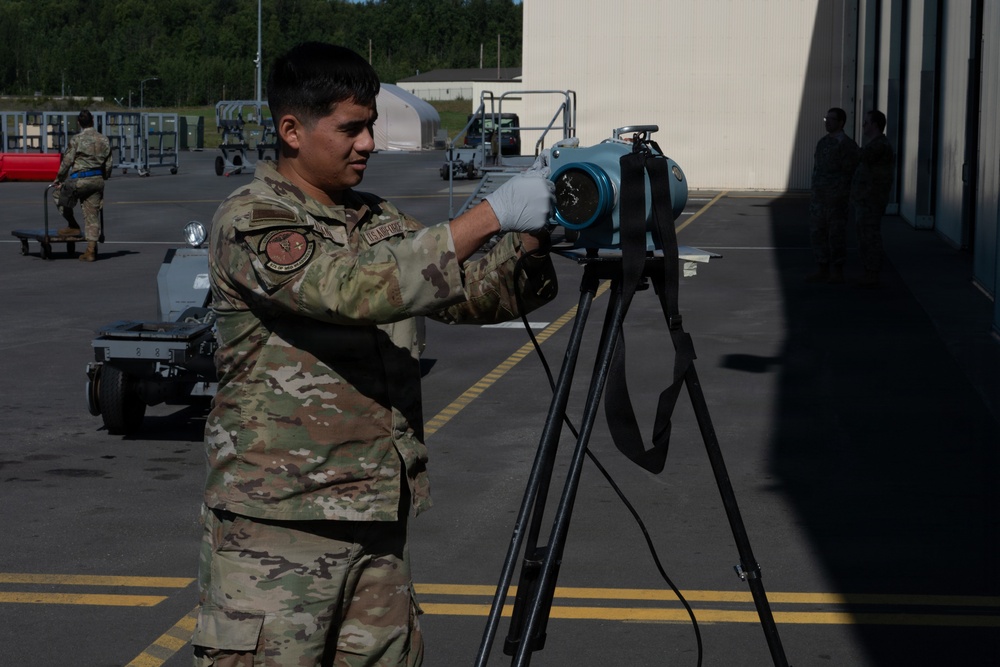A U.S. Air Force service member in OCP (Operational Camouflage Pattern) uniform is operating or adjusting a blue cylindrical device mounted on a tripod on an airfield tarmac. The setting appears to be a military air base with hangars, ground support equipment, and other personnel visible in the background. The activity suggests equipment setup or calibration operations.