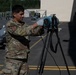 A U.S. Air Force service member in OCP (Operational Camouflage Pattern) uniform is operating or adjusting a blue cylindrical device mounted on a tripod on an airfield tarmac. The setting appears to be a military air base with hangars, ground support equipment, and other personnel visible in the background. The activity suggests equipment setup or calibration operations.