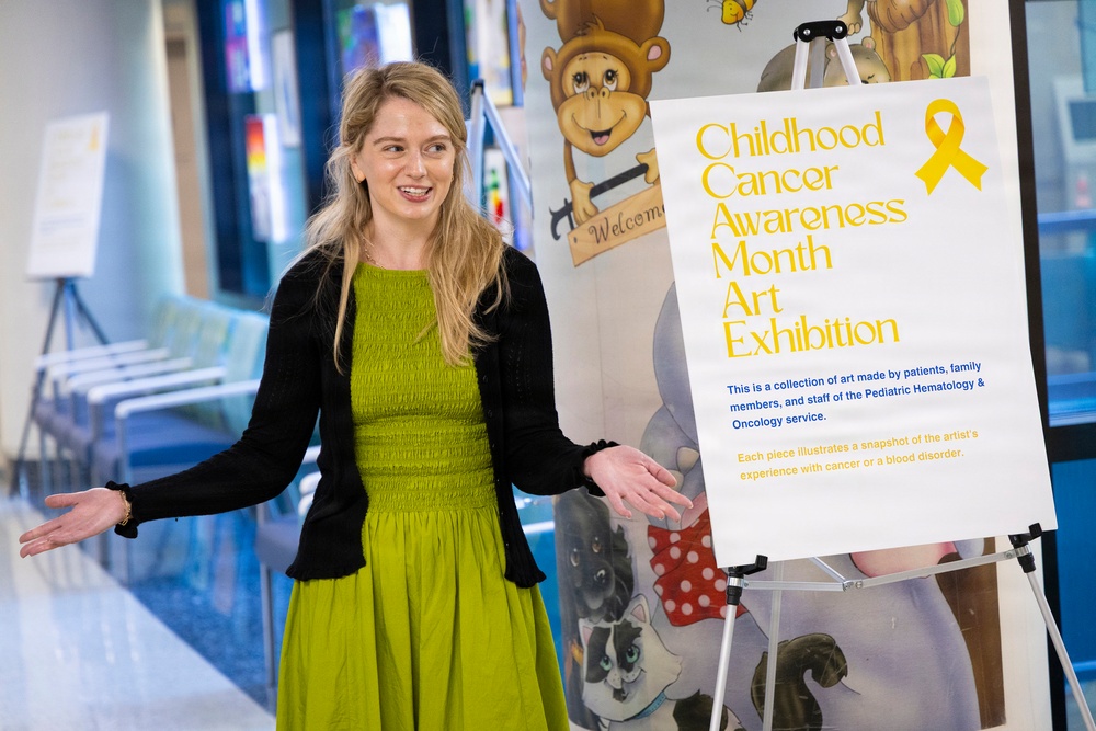A woman with blonde hair wearing a green dress and black cardigan stands next to a display sign for a 'Childhood Cancer Awareness Month Art Exhibition' in what appears to be a hospital or medical facility corridor. She is gesturing with both arms outstretched in a welcoming or presenting manner. The exhibition features artwork made by patients, family members, and staff of a Pediatric Hematology &amp; Oncology service.