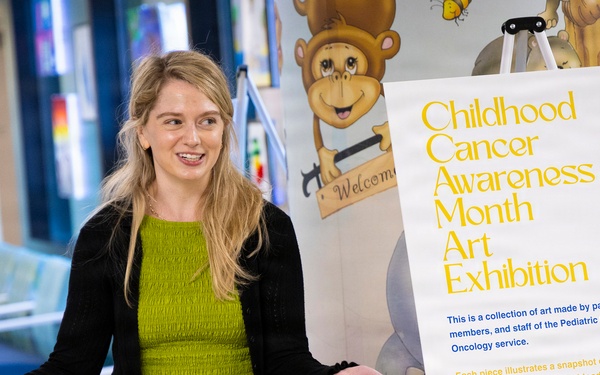 A woman with blonde hair wearing a green dress and black cardigan stands next to a display sign for a 'Childhood Cancer Awareness Month Art Exhibition' in what appears to be a hospital or medical facility corridor. She is gesturing with both arms outstretched in a welcoming or presenting manner. The exhibition features artwork made by patients, family members, and staff of a Pediatric Hematology &amp; Oncology service.