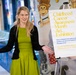 A woman with blonde hair wearing a green dress and black cardigan stands next to a display sign for a 'Childhood Cancer Awareness Month Art Exhibition' in what appears to be a hospital or medical facility corridor. She is gesturing with both arms outstretched in a welcoming or presenting manner. The exhibition features artwork made by patients, family members, and staff of a Pediatric Hematology &amp; Oncology service.