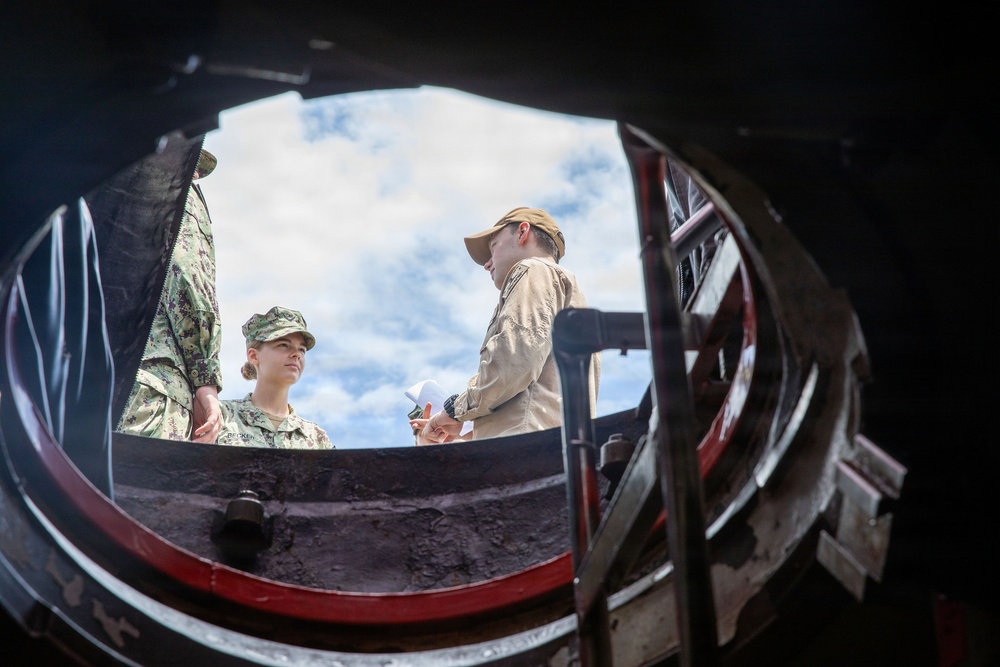 The image is taken from below looking up through a circular hatch or opening, showing two individuals standing above it against a partly cloudy sky. One person is wearing military camouflage uniform with a patrol cap, while the other is in civilian tan/khaki clothing and a baseball cap. The perspective creates a dramatic fisheye-like framing effect through the dark circular structure.