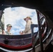 The image is taken from below looking up through a circular hatch or opening, showing two individuals standing above it against a partly cloudy sky. One person is wearing military camouflage uniform with a patrol cap, while the other is in civilian tan/khaki clothing and a baseball cap. The perspective creates a dramatic fisheye-like framing effect through the dark circular structure.