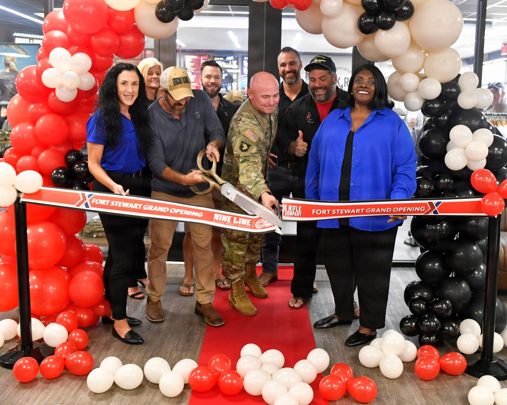 A grand opening ribbon-cutting ceremony is taking place at Fort Stewart for a Nine Line store, also featuring Oak Rifle Company branding. A group of approximately eight people, including a U.S. Army soldier in camouflage uniform, are gathered around a large ceremonial ribbon using oversized golden scissors. The event is decorated with red, black, and white/cream balloon arches and a red carpet.
