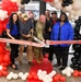 A grand opening ribbon-cutting ceremony is taking place at Fort Stewart for a Nine Line store, also featuring Oak Rifle Company branding. A group of approximately eight people, including a U.S. Army soldier in camouflage uniform, are gathered around a large ceremonial ribbon using oversized golden scissors. The event is decorated with red, black, and white/cream balloon arches and a red carpet.