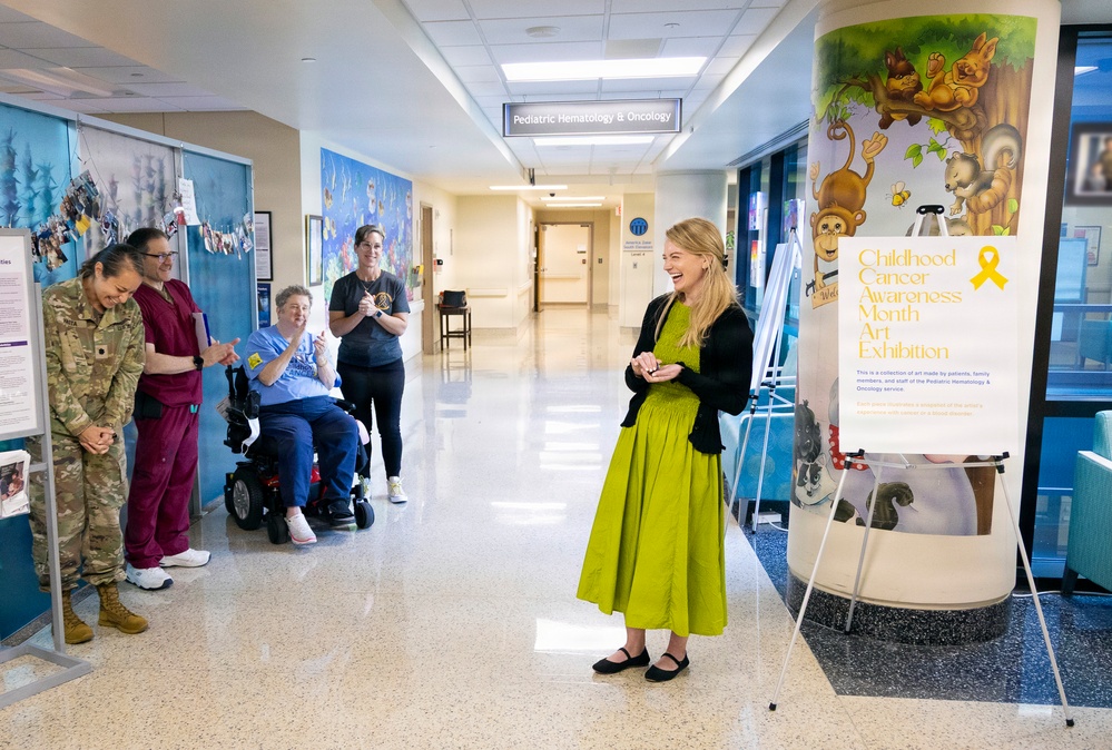 A woman in a green dress stands smiling in the hallway of a Pediatric Hematology &amp; Oncology unit, facing a small group of hospital staff who are applauding her. The setting appears to be a formal or celebratory moment, possibly a recognition event or visit, near a 'Childhood Cancer Awareness Month Art Exhibition' display. The hallway features colorful murals and medical facility signage.