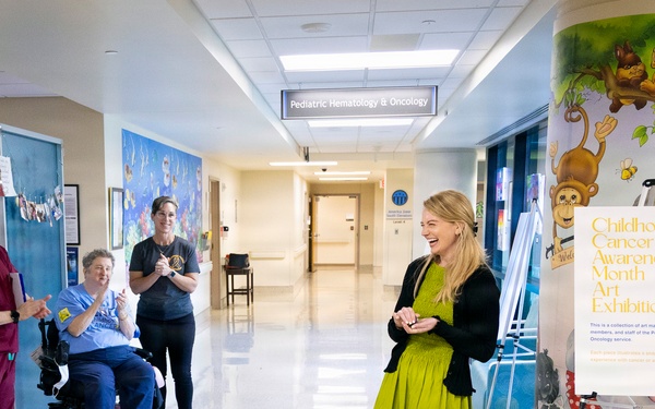 A woman in a green dress stands smiling in the hallway of a Pediatric Hematology &amp; Oncology unit, facing a small group of hospital staff who are applauding her. The setting appears to be a formal or celebratory moment, possibly a recognition event or visit, near a 'Childhood Cancer Awareness Month Art Exhibition' display. The hallway features colorful murals and medical facility signage.
