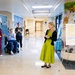 A woman in a green dress stands smiling in the hallway of a Pediatric Hematology &amp; Oncology unit, facing a small group of hospital staff who are applauding her. The setting appears to be a formal or celebratory moment, possibly a recognition event or visit, near a 'Childhood Cancer Awareness Month Art Exhibition' display. The hallway features colorful murals and medical facility signage.