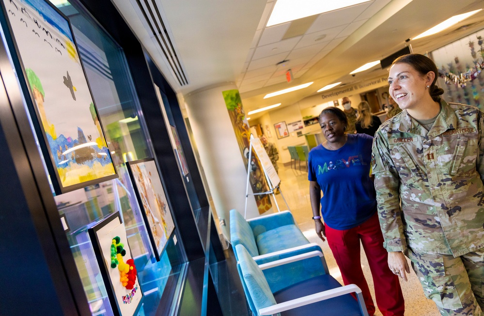 A U.S. Air Force service member, identified by name tape as 'QUILLEN,' and a civilian woman in a blue Marvel t-shirt are viewing framed children's artwork displayed on a wall in what appears to be a medical facility hallway. The setting appears to be a pediatric or developmental/behavioral health clinic, as indicated by signage visible in the background. Both individuals appear engaged and smiling while observing the artwork display.