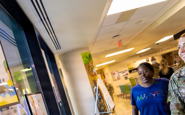 A U.S. Air Force service member, identified by name tape as 'QUILLEN,' and a civilian woman in a blue Marvel t-shirt are viewing framed children's artwork displayed on a wall in what appears to be a medical facility hallway. The setting appears to be a pediatric or developmental/behavioral health clinic, as indicated by signage visible in the background. Both individuals appear engaged and smiling while observing the artwork display.