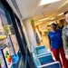 A U.S. Air Force service member, identified by name tape as 'QUILLEN,' and a civilian woman in a blue Marvel t-shirt are viewing framed children's artwork displayed on a wall in what appears to be a medical facility hallway. The setting appears to be a pediatric or developmental/behavioral health clinic, as indicated by signage visible in the background. Both individuals appear engaged and smiling while observing the artwork display.