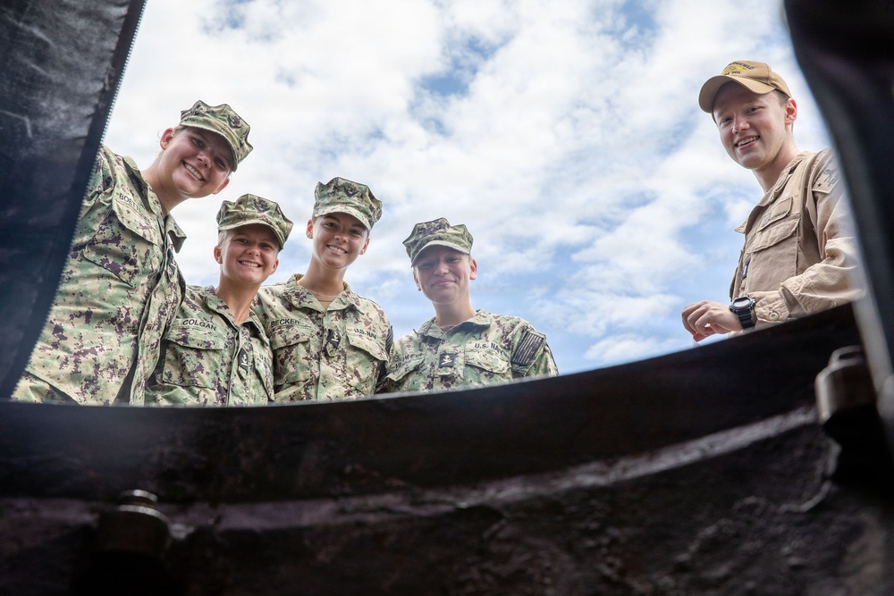 Five military personnel, four in U.S. Navy digital camouflage uniforms and one in tan/khaki uniform, are photographed from a low-angle perspective looking down into what appears to be a large container or vessel. The photo is taken from below, creating a dramatic upward perspective with a partly cloudy sky in the background. Visible name tapes include 'COLGAN' and 'BECKER' on two of the individuals.