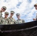 Five military personnel, four in U.S. Navy digital camouflage uniforms and one in tan/khaki uniform, are photographed from a low-angle perspective looking down into what appears to be a large container or vessel. The photo is taken from below, creating a dramatic upward perspective with a partly cloudy sky in the background. Visible name tapes include 'COLGAN' and 'BECKER' on two of the individuals.