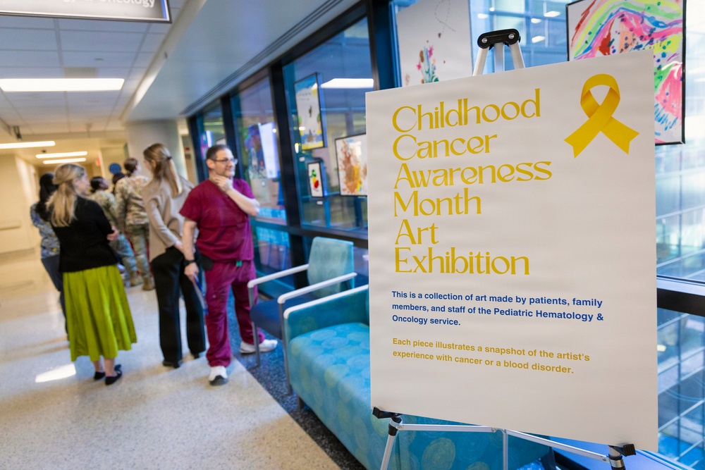 The image shows a Childhood Cancer Awareness Month Art Exhibition displayed in what appears to be a hospital corridor or lobby. A prominent sign on an easel in the foreground announces the exhibition, which features artwork created by patients, family members, and staff of the Pediatric Hematology &amp; Oncology service. Several people, including a medical professional in burgundy scrubs, are gathered in the background conversing near the exhibition.