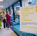 The image shows a Childhood Cancer Awareness Month Art Exhibition displayed in what appears to be a hospital corridor or lobby. A prominent sign on an easel in the foreground announces the exhibition, which features artwork created by patients, family members, and staff of the Pediatric Hematology &amp; Oncology service. Several people, including a medical professional in burgundy scrubs, are gathered in the background conversing near the exhibition.