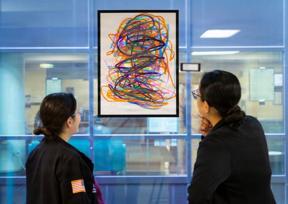 Two individuals wearing dark jackets are viewing a framed abstract artwork displayed on a glass wall or window. The artwork features colorful, overlapping scribble-like lines in multiple colors including orange, purple, blue, pink, and green on a white background. The setting appears to be an institutional or public building interior.