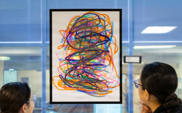 Two individuals wearing dark jackets are viewing a framed abstract artwork displayed on a glass wall or window. The artwork features colorful, overlapping scribble-like lines in multiple colors including orange, purple, blue, pink, and green on a white background. The setting appears to be an institutional or public building interior.