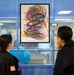 Two individuals wearing dark jackets are viewing a framed abstract artwork displayed on a glass wall or window. The artwork features colorful, overlapping scribble-like lines in multiple colors including orange, purple, blue, pink, and green on a white background. The setting appears to be an institutional or public building interior.