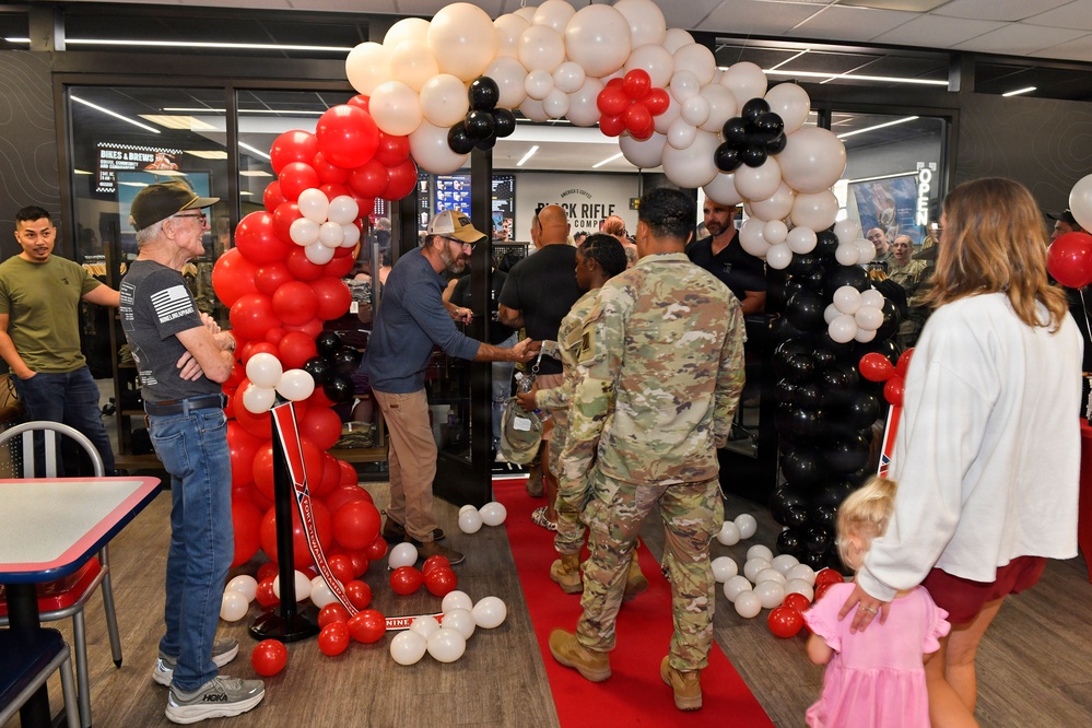 A grand opening or ribbon-cutting ceremony for a Black Rifle Coffee Company location, featuring a decorative balloon arch in red, white, and black colors over a red carpet entrance. Military personnel in camouflage uniforms are present alongside civilians, with handshakes being exchanged near the entrance. The event appears festive and celebratory with scattered balloons on the floor.