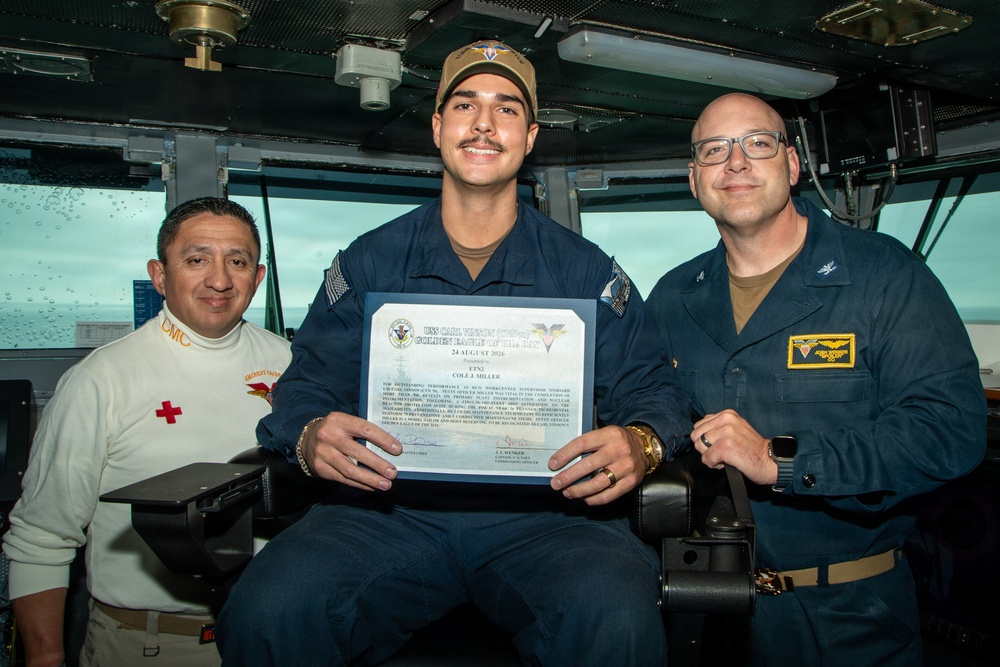 Three individuals pose for a photo aboard what appears to be a U.S. Navy vessel's bridge or command area. The central figure is holding a certificate titled 'Golden Eagle of the Day' dated 24 August 2026, presented to 'ET52 Cole J. Miller' from the USS Carl Vinson. The occasion appears to be a military recognition ceremony.