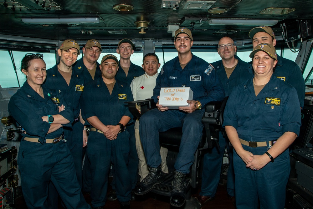 A group of approximately eleven U.S. Navy personnel are posed together on what appears to be a ship's bridge or command center. One central figure is seated and holding a white box with the text 'GOLD EAGLE OF THE DAY' written on it, suggesting a recognition or award ceremony. The group includes both male and female sailors in standard Navy blue coveralls, with one individual wearing a white uniform with a red cross insignia.