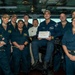 A group of approximately eleven U.S. Navy personnel are posed together on what appears to be a ship's bridge or command center. One central figure is seated and holding a white box with the text 'GOLD EAGLE OF THE DAY' written on it, suggesting a recognition or award ceremony. The group includes both male and female sailors in standard Navy blue coveralls, with one individual wearing a white uniform with a red cross insignia.