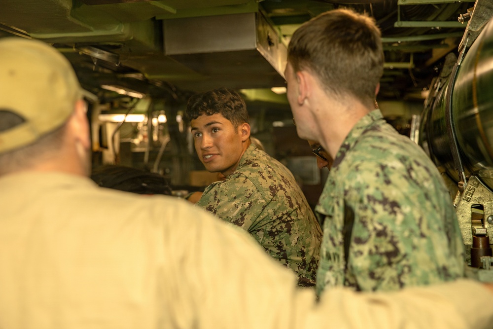 The image shows military personnel in digital camouflage uniforms gathered in what appears to be a confined mechanical or engineering space, likely aboard a naval vessel or inside a military vehicle. At least three individuals are visible, with one facing the camera appearing to speak or explain something. The setting is characterized by tight quarters with visible machinery, pipes, and equipment.