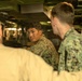 The image shows military personnel in digital camouflage uniforms gathered in what appears to be a confined mechanical or engineering space, likely aboard a naval vessel or inside a military vehicle. At least three individuals are visible, with one facing the camera appearing to speak or explain something. The setting is characterized by tight quarters with visible machinery, pipes, and equipment.