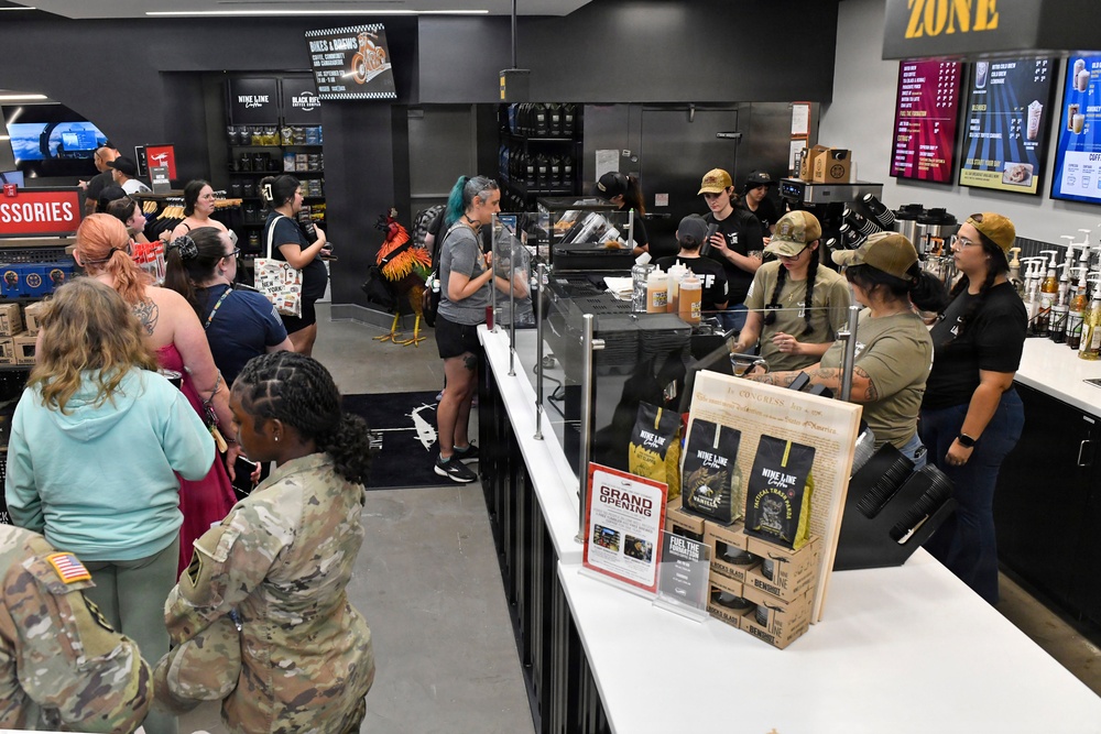 A busy grand opening event at a Nine Line Coffee shop, likely located on or near a military installation, evidenced by the presence of uniformed military personnel and patriotic branding. Customers are lined up along a white counter being served by multiple staff members in matching attire. The store features coffee merchandise, digital menu boards, and patriotic/military-themed decor.