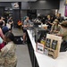 A busy grand opening event at a Nine Line Coffee shop, likely located on or near a military installation, evidenced by the presence of uniformed military personnel and patriotic branding. Customers are lined up along a white counter being served by multiple staff members in matching attire. The store features coffee merchandise, digital menu boards, and patriotic/military-themed decor.