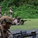 Military personnel in camouflage uniforms are conducting a live-fire or marksmanship training exercise in a tropical outdoor setting. Several soldiers are in prone firing positions with rifles aimed downrange, while others stand observing or supervising. The lush green vegetation in the background suggests a tropical or subtropical training environment.