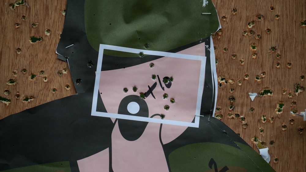 The image shows a damaged poster or billboard lying on a wooden surface, riddled with numerous bullet holes. The poster appears to feature a stylized graphic design with pink, black, and dark green colors, along with what appears to be Arabic or Persian script. The extensive bullet damage to both the poster and the underlying wooden surface indicates deliberate or combat-related destruction.