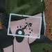 The image shows a damaged poster or billboard lying on a wooden surface, riddled with numerous bullet holes. The poster appears to feature a stylized graphic design with pink, black, and dark green colors, along with what appears to be Arabic or Persian script. The extensive bullet damage to both the poster and the underlying wooden surface indicates deliberate or combat-related destruction.