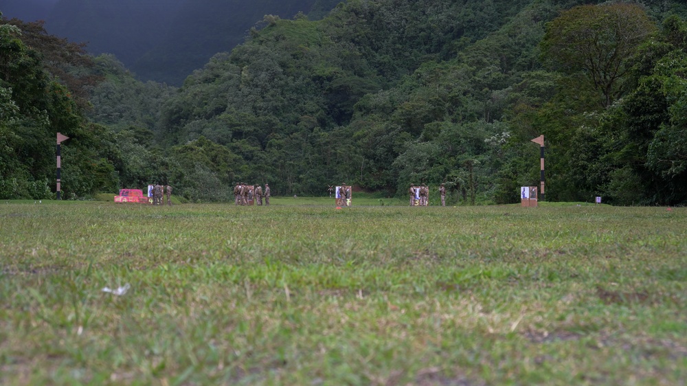 A wide grass field serves as what appears to be a military shooting range or training area, with personnel in camouflage uniforms gathered at the far end near target stands. The setting is surrounded by dense tropical or subtropical forest with lush green vegetation and hills in the background. The photo is taken from ground level, emphasizing the expanse of the field.