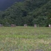 A wide grass field serves as what appears to be a military shooting range or training area, with personnel in camouflage uniforms gathered at the far end near target stands. The setting is surrounded by dense tropical or subtropical forest with lush green vegetation and hills in the background. The photo is taken from ground level, emphasizing the expanse of the field.