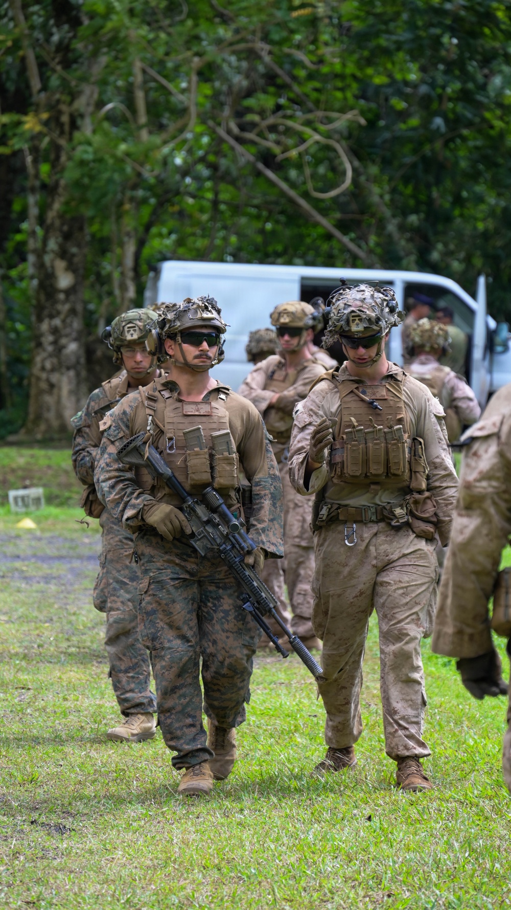 A group of military personnel in full combat gear are walking across a grassy area, likely during a training exercise or field operation. The soldiers are equipped with tactical vests, helmets with camouflage netting, weapons, and protective eyewear. The setting appears to be a tropical or subtropical outdoor environment with dense green vegetation in the background.