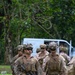 A group of military personnel in full combat gear are walking across a grassy area, likely during a training exercise or field operation. The soldiers are equipped with tactical vests, helmets with camouflage netting, weapons, and protective eyewear. The setting appears to be a tropical or subtropical outdoor environment with dense green vegetation in the background.