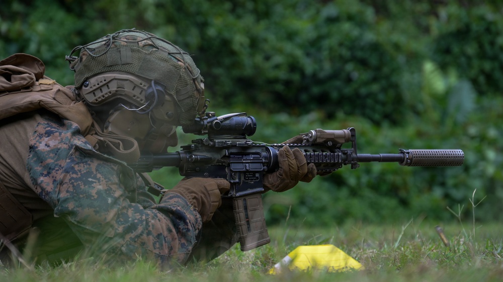 A military service member in MARPAT (Marine pattern) digital camouflage uniform is in a prone firing position on grass, aiming a rifle downrange. The soldier is equipped with full tactical gear including a combat helmet with rail accessories, plate carrier, and tactical gloves. The setting appears to be an outdoor training range or field exercise environment with dense green vegetation in the background.