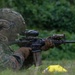 A military service member in MARPAT (Marine pattern) digital camouflage uniform is in a prone firing position on grass, aiming a rifle downrange. The soldier is equipped with full tactical gear including a combat helmet with rail accessories, plate carrier, and tactical gloves. The setting appears to be an outdoor training range or field exercise environment with dense green vegetation in the background.