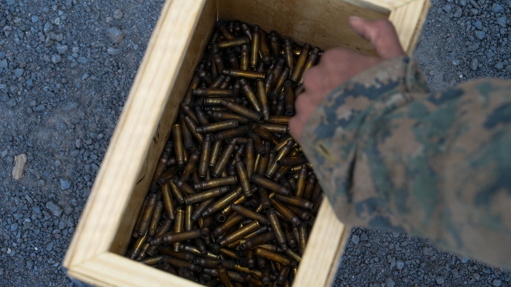 A top-down view of a wooden crate filled with numerous brass cartridge casings, likely spent shell casings collected after a firing exercise. A person wearing military camouflage uniform (consistent with USMC MARPAT pattern) is reaching into or over the crate. The crate is resting on a gravel surface, suggesting an outdoor military range environment.