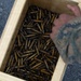 A top-down view of a wooden crate filled with numerous brass cartridge casings, likely spent shell casings collected after a firing exercise. A person wearing military camouflage uniform (consistent with USMC MARPAT pattern) is reaching into or over the crate. The crate is resting on a gravel surface, suggesting an outdoor military range environment.