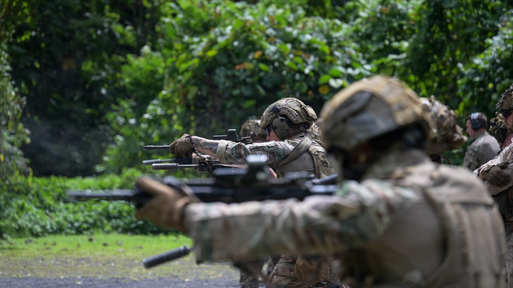 A line of military personnel in full combat gear are engaged in a live-fire shooting range exercise. Soldiers are wearing multicam/camouflage uniforms, tactical helmets, and hearing protection while aiming rifles downrange. The setting appears to be an outdoor range bordered by dense tropical or subtropical vegetation.