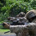 A line of military personnel in full combat gear are engaged in a live-fire shooting range exercise. Soldiers are wearing multicam/camouflage uniforms, tactical helmets, and hearing protection while aiming rifles downrange. The setting appears to be an outdoor range bordered by dense tropical or subtropical vegetation.