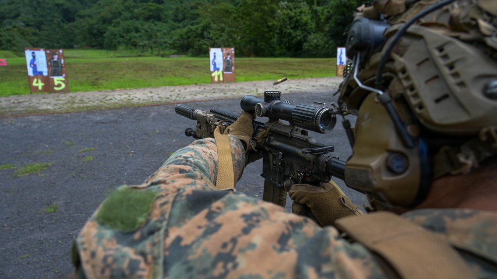 A military service member in MARPAT (Marine pattern) camouflage uniform is aiming a rifle equipped with a magnified optic scope at a shooting range. Multiple numbered paper silhouette targets are visible downrange on a gravel surface backed by dense green vegetation. The image depicts a live-fire training exercise at an outdoor range.