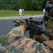 A military service member in MARPAT (Marine pattern) camouflage uniform is aiming a rifle equipped with a magnified optic scope at a shooting range. Multiple numbered paper silhouette targets are visible downrange on a gravel surface backed by dense green vegetation. The image depicts a live-fire training exercise at an outdoor range.
