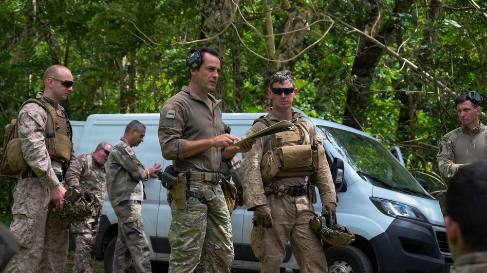 A group of military or tactical personnel in various camouflage uniforms are gathered in a wooded outdoor area near a white van. Two central figures appear to be reviewing a document or map together, suggesting a briefing or planning session. The personnel are equipped with tactical vests, pouches, and sidearms, indicating an operational or training exercise.
